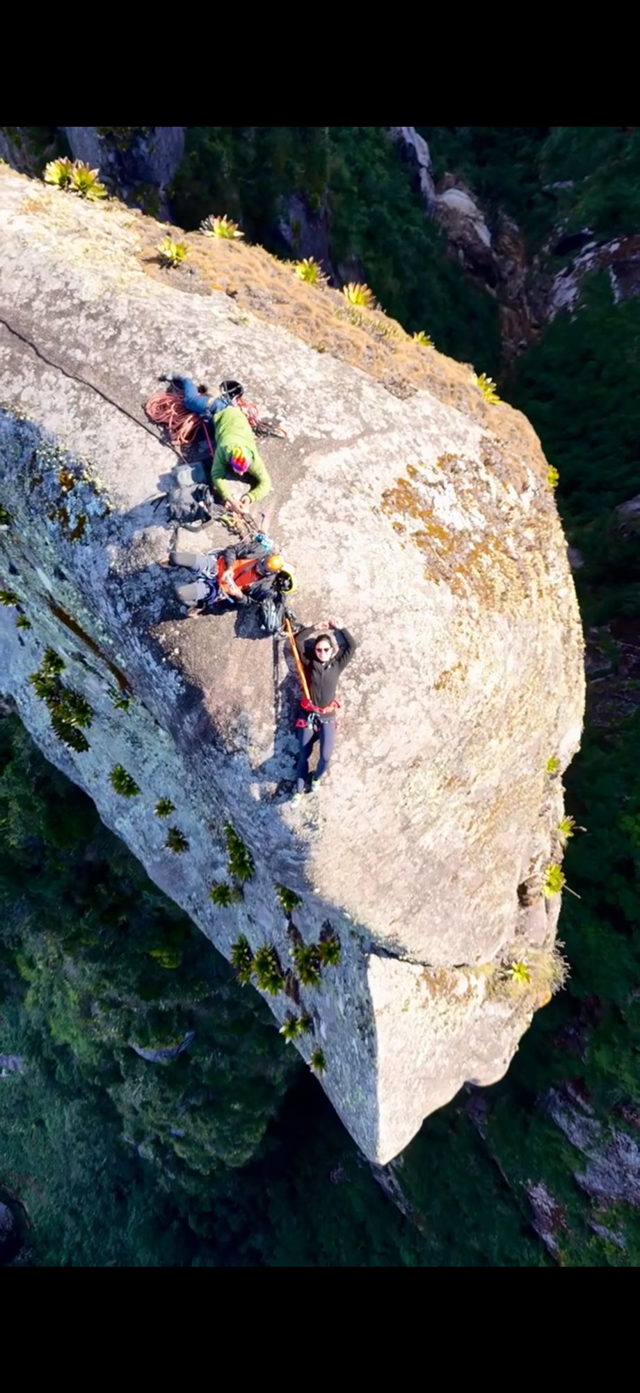 Vista aérea de três escaladores em parede inclinada da Agulha do Diabo com floresta densa e vazio abaixo