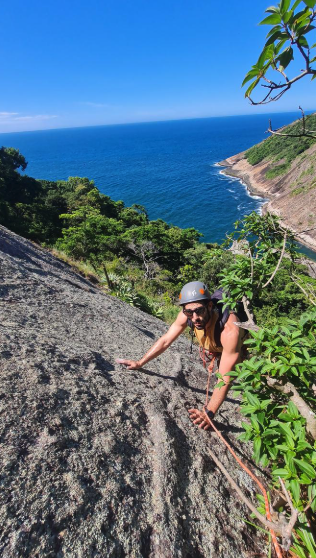 Escalador com capacete em placa de granito da Agulha Guarischi com o mar azul de Niterói ao fundo
