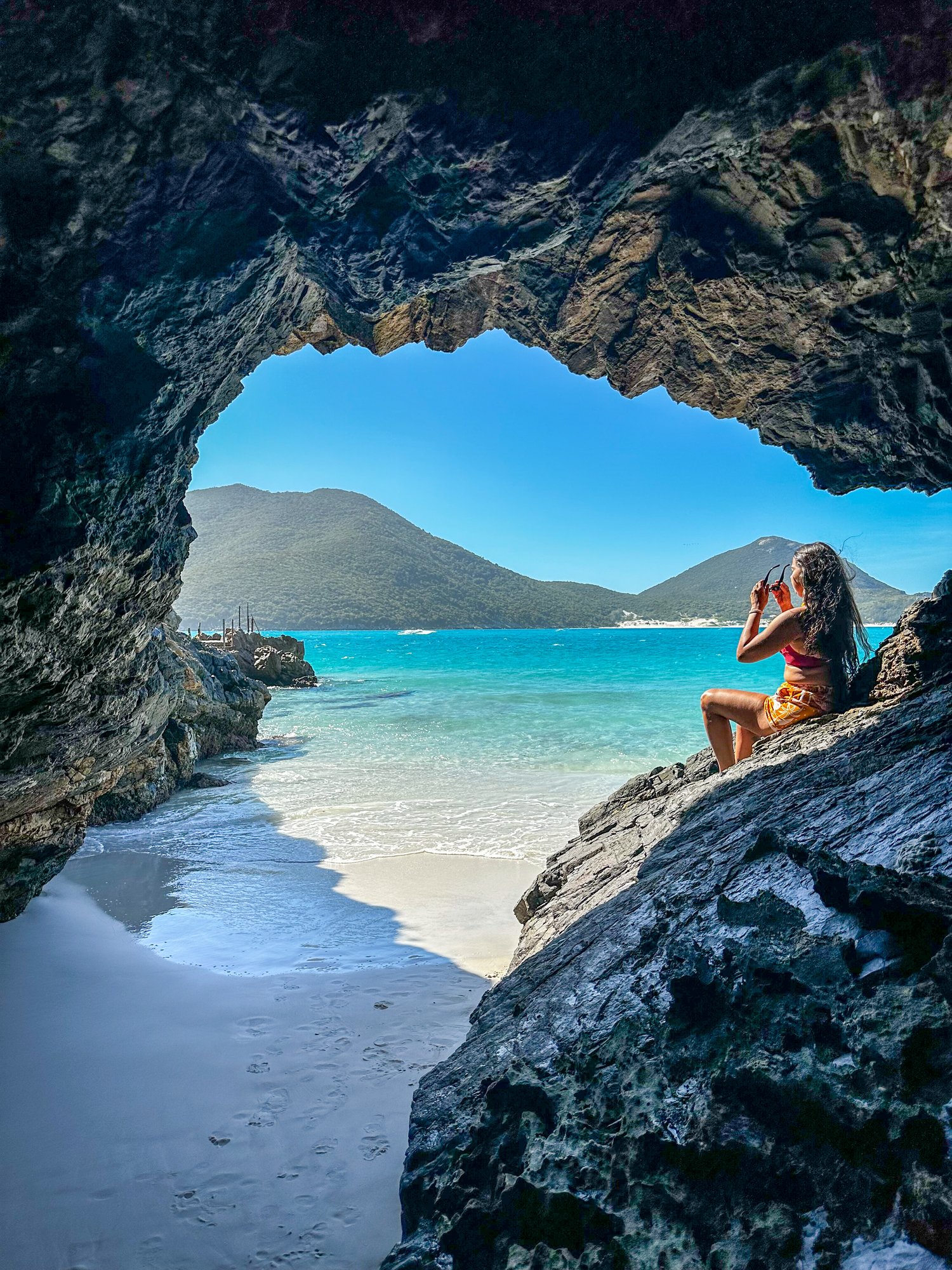 Mulher sentada em arco de rocha natural com praia paradisíaca ao fundo