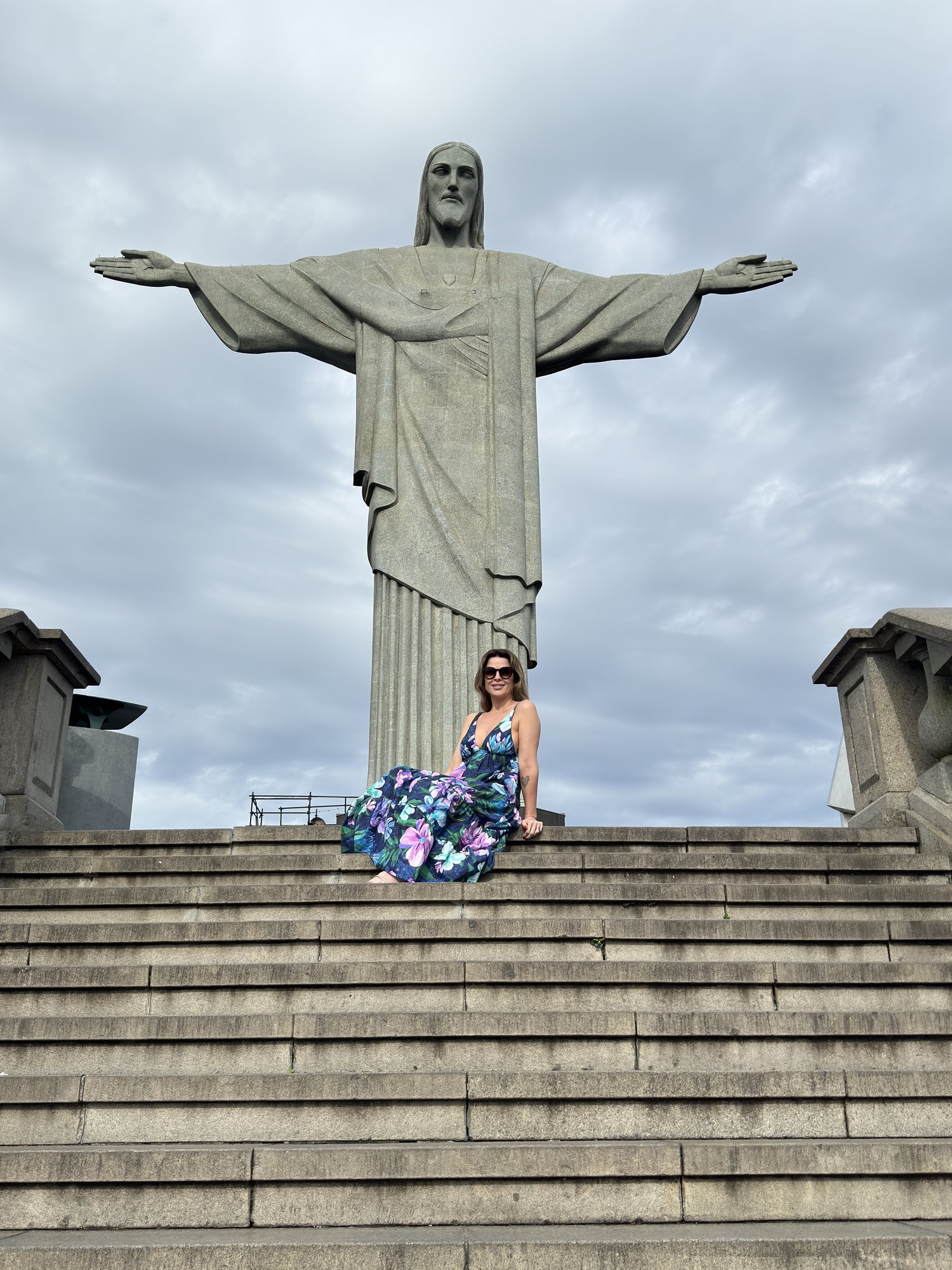 Visitante com vestido floral sentada nos degraus do Cristo Redentor com o Rio de Janeiro abaixo
