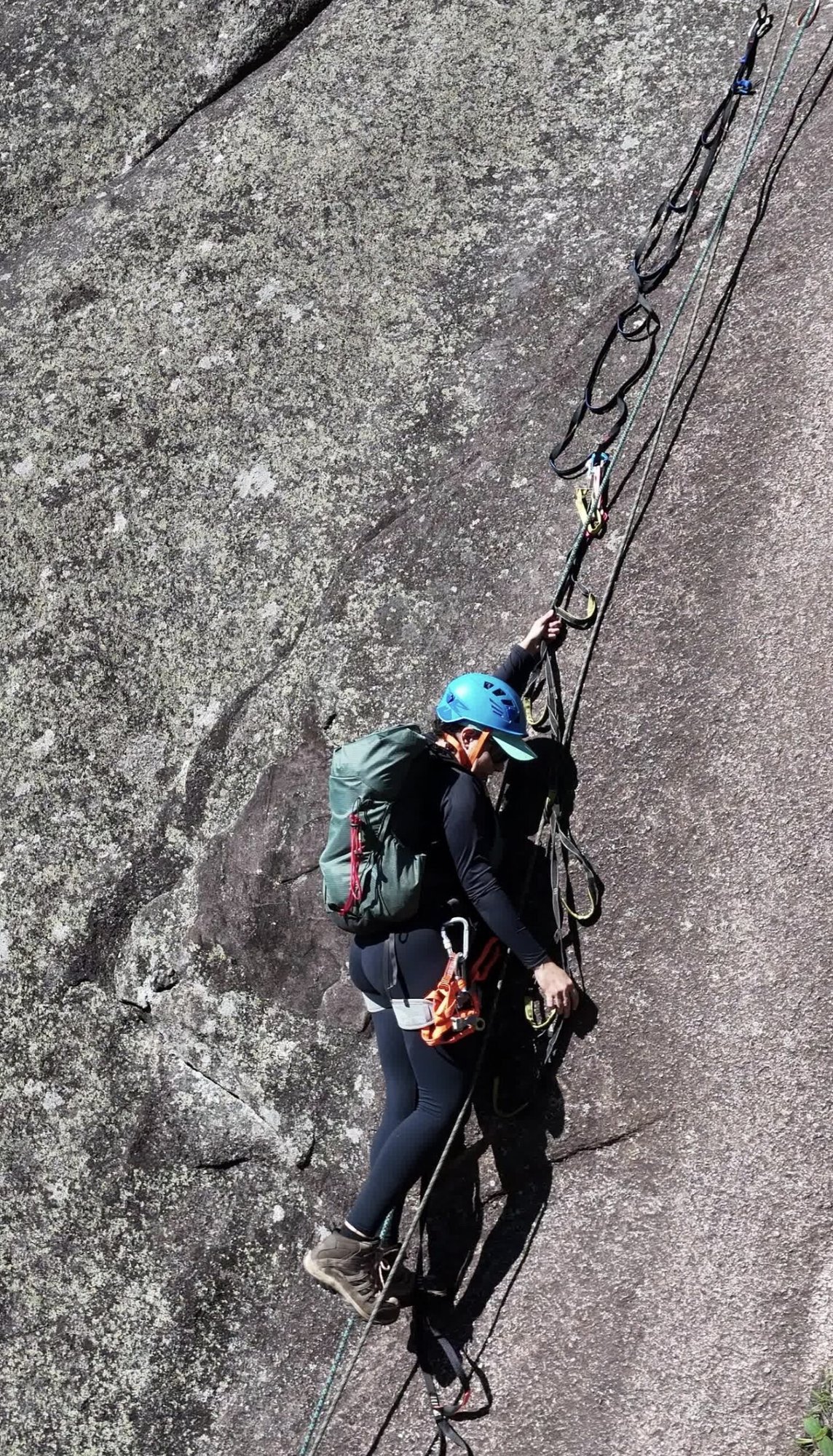 Escaladora com capacete azul e mochila verde em placa de granito no Dedo de Nossa Senhora ajustando equipamento