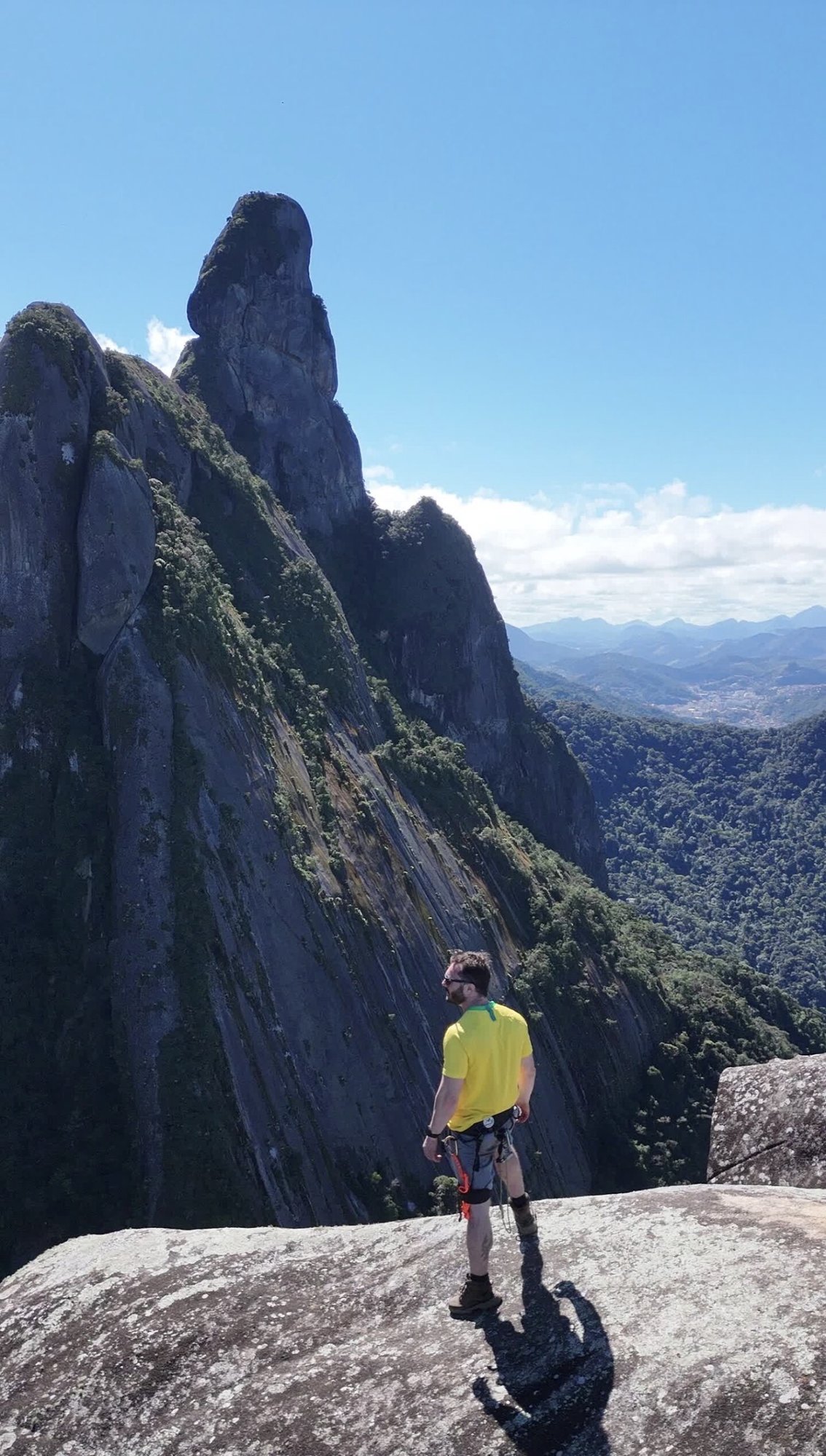 Escalador de camiseta amarela em patamar rochoso com o Dedo de Nossa Senhora em destaque e Teresópolis ao fundo no vale