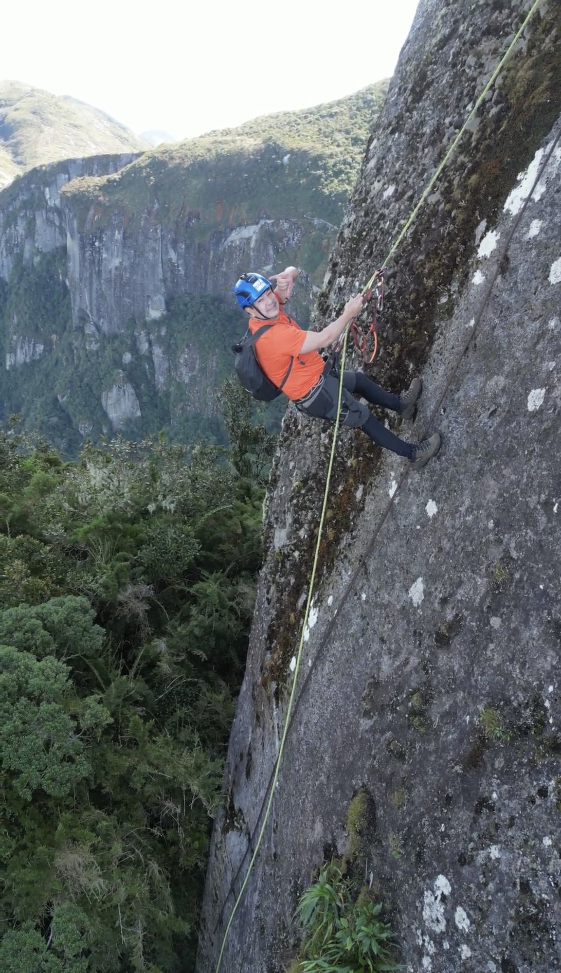 Escalador de capacete azul e camiseta laranja em fissura vertical no Garrafão com paredões de granito ao fundo