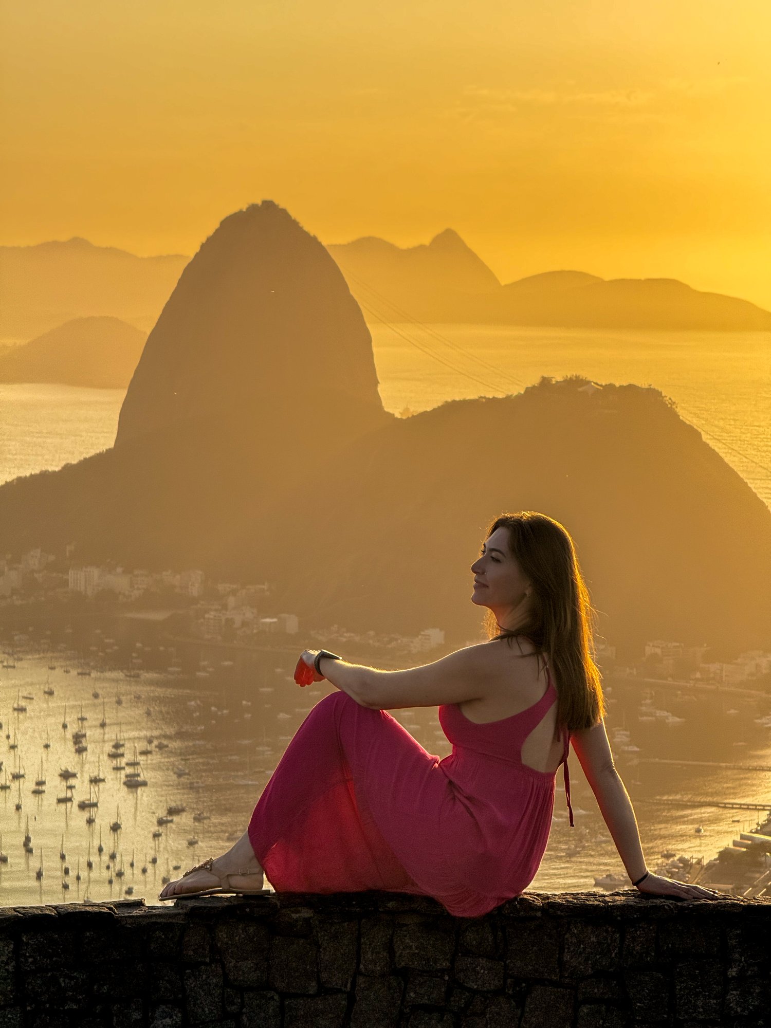 Visitante com vestido rosa sentada no muro do Mirante Dona Marta admirando o Pão de Açúcar na luz dourada do amanhecer