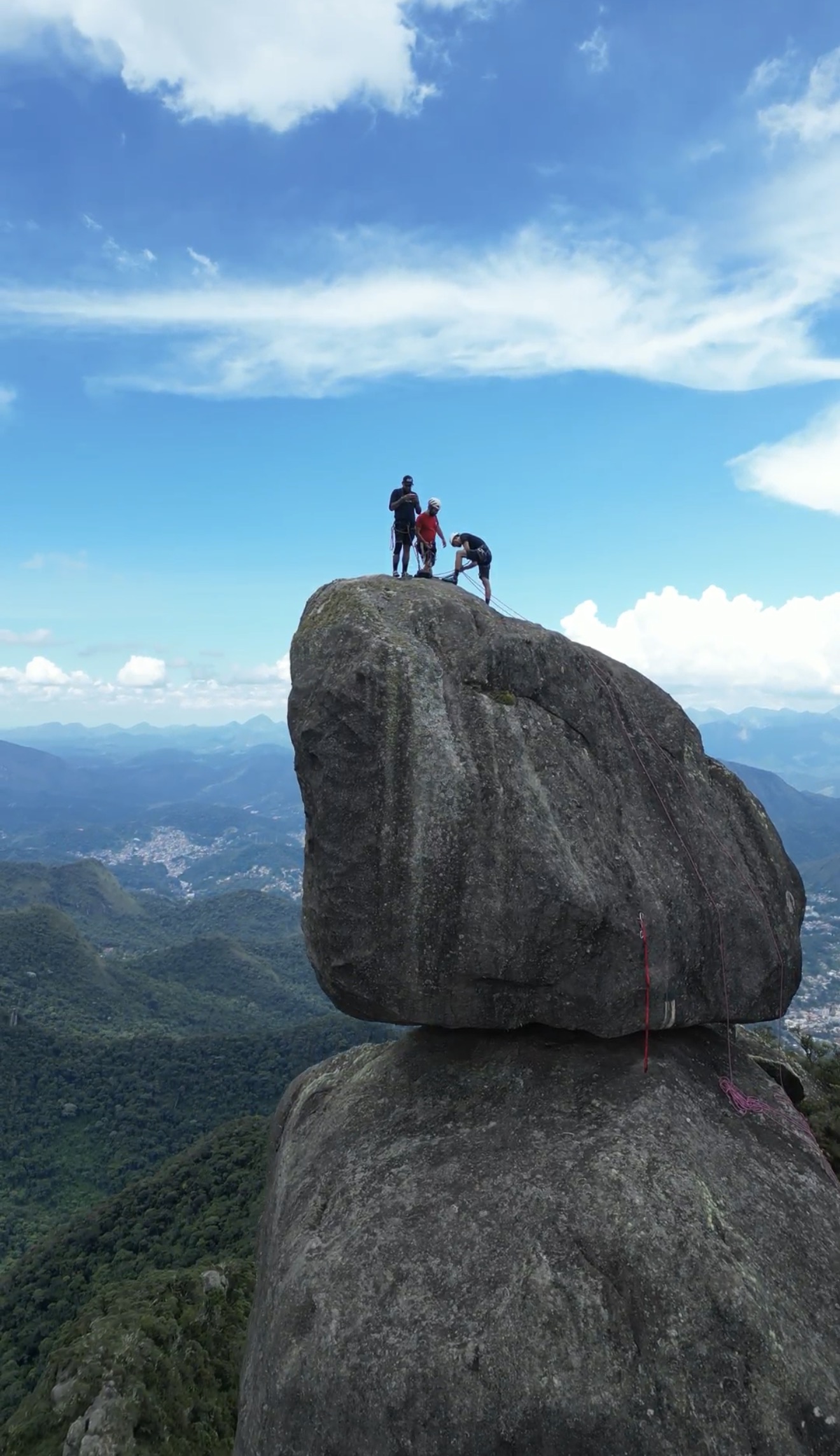 Três escaladores no topo do bloco da Verruga do Frade com corda vermelha e panorama amplo do vale de Teresópolis
